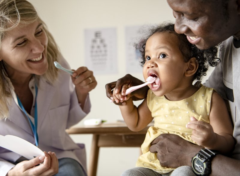 a dentist holding a toothbrush and smiling while a father holds his baby and helps her brush her teeth